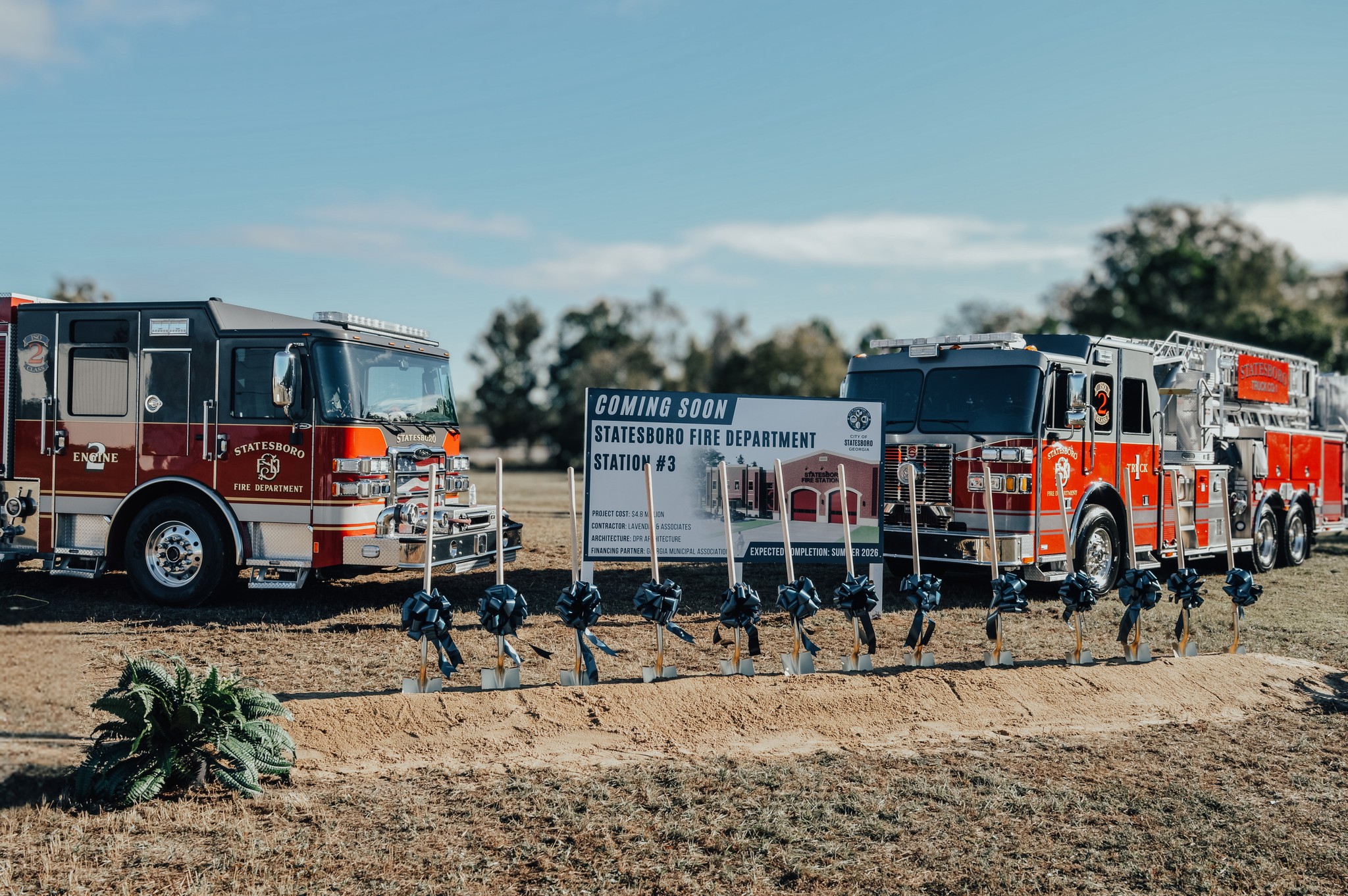 two fire engines parked around a "coming soon" sign featuring the design for the new fire department with a line of golden shovels with navy blue bows for the ground breaking ceremony